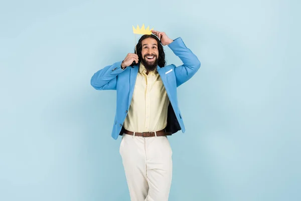 Щасливий іспаномовний чоловік в куртці тримає паперову корону на паличці ізольовано на синьому — стокове фото