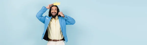 Glücklicher hispanischer Mann in Jacke mit Papierkrone auf Stick isoliert auf blauem Banner — Stockfoto