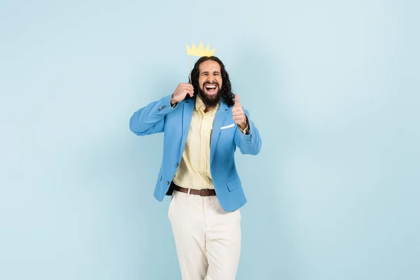 Homem hispânico feliz na jaqueta segurando coroa de papel na vara e mostrando polegar para cima isolado no azul — Fotografia de Stock