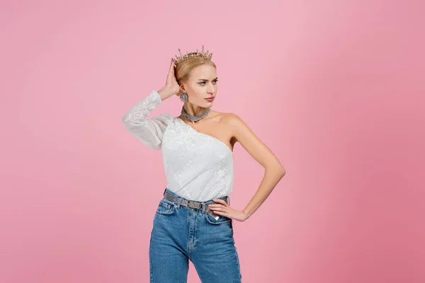 Mujer rubia ajustando la corona de lujo y de pie con la mano en la cadera aislada en rosa — Stock Photo