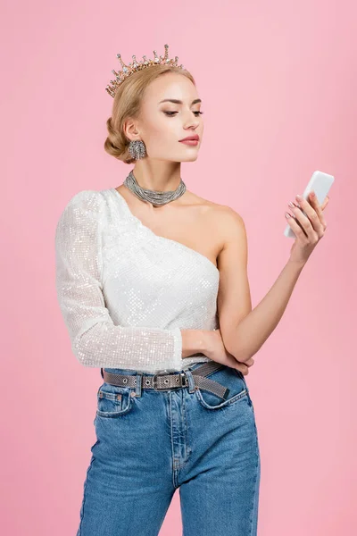 Блондинка в розкішній короні дивиться на смартфон ізольовано на рожевому — стокове фото
