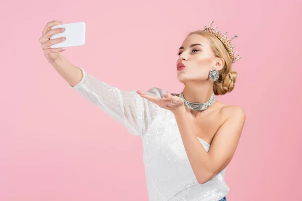 Mulher loira em coroa de luxo tomando selfie no smartphone e enviando beijo de ar isolado no rosa — Fotografia de Stock