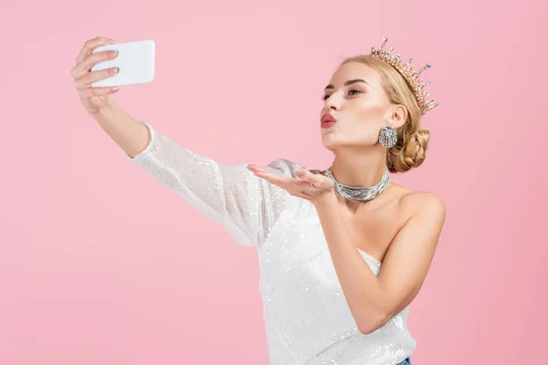 Mujer rubia en la corona de lujo tomando selfie en el teléfono inteligente y el envío de beso de aire aislado en rosa — Stock Photo