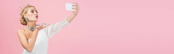 Mujer rubia en la corona de lujo tomando selfie en el teléfono inteligente y el envío de beso de aire aislado en rosa, bandera — Stock Photo