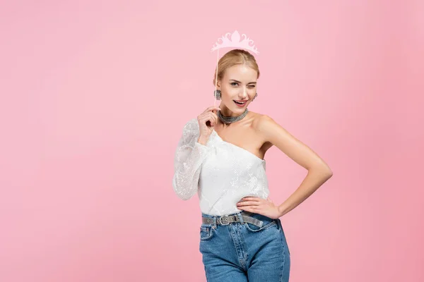 Mujer rubia sosteniendo la corona de papel en palo y de pie con la mano en la cadera aislado en rosa — Stock Photo