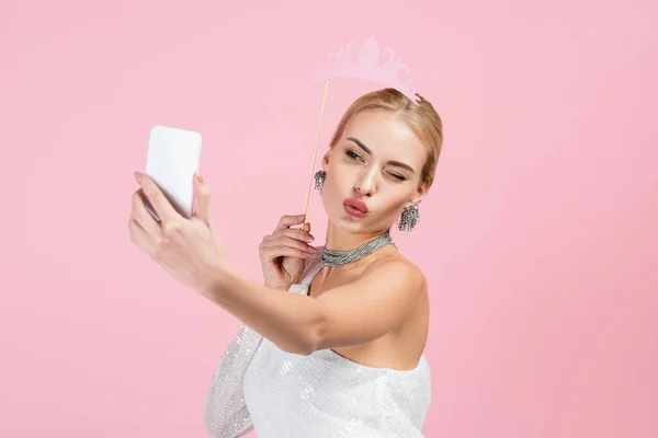 Блондинка тримає паперову корону на паличці і приймає селфі, посилаючи повітряний поцілунок ізольовано на рожевому — стокове фото