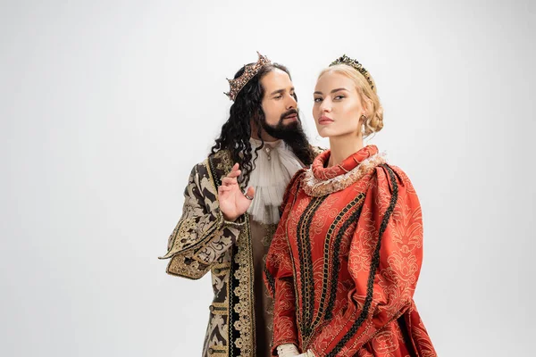 Rei hispânico em coroa e roupas medievais olhando para a esposa loira isolada no branco — Fotografia de Stock