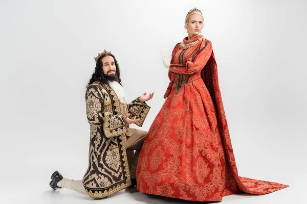 Латиноамериканский король в короне и средневековой одежде, стоящий на коленях, в то время как блондинка королева не показывает жеста на белом — стоковое фото