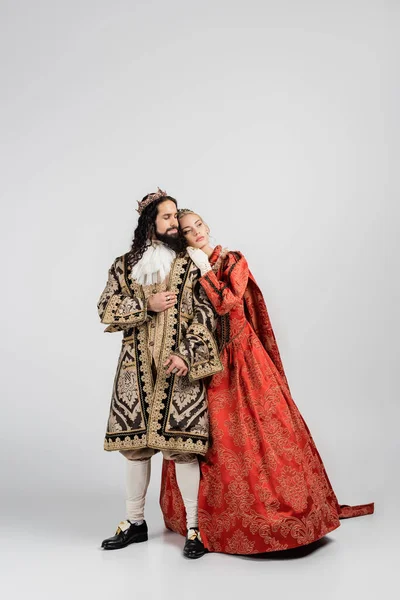 Longitud completa de la reina rubia en corona real apoyada en el rey hispano en ropa medieval en blanco - foto de stock