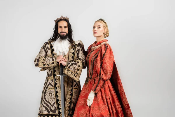 Межрасовая историческая пара в средневековой одежде и коронах, смотрящая на камеру, изолированную на белом — стоковое фото