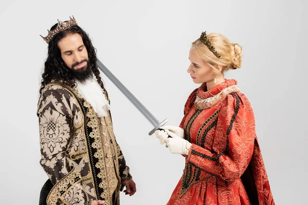 Reina rubia sosteniendo espada cerca del cuello del rey hispano en ropa medieval aislada en blanco - foto de stock