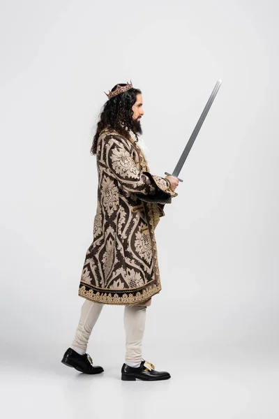 Side view of hispanic king in medieval clothing and crown holding sword on white — Stock Photo