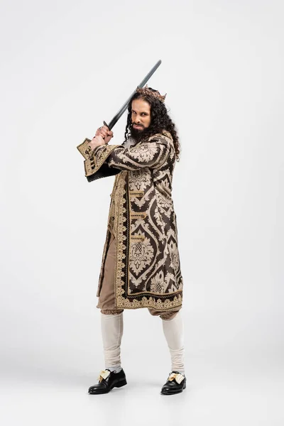 Full length of hispanic king in crown and medieval clothing holding sword on white — Stock Photo