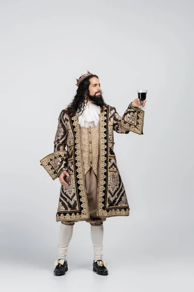 Full length of hispanic king in medieval clothing and crown holding glass of red wine on white — Stock Photo