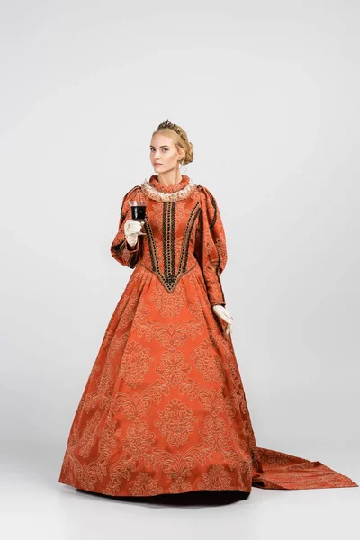 Full length of queen in dress and crown holding glass of red wine on white — Stock Photo