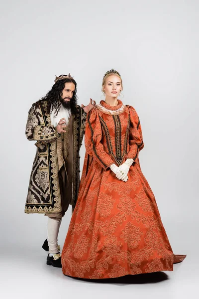 Longitud completa del rey hispano en ropa medieval de pie detrás de la reina en la corona en blanco - foto de stock