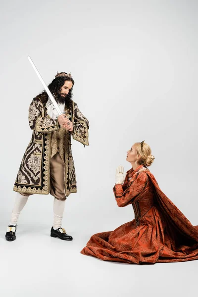 Full length of cruel hispanic king in medieval clothing holding sword near scared queen in crown sitting with praying hands on white — Stock Photo