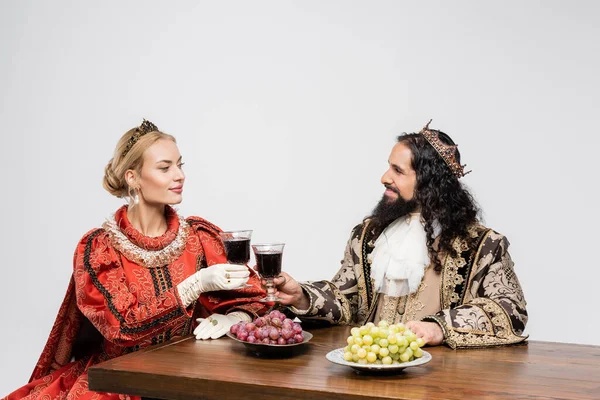 Interracial historical couple in crowns clinking glasses of red wine while sitting at table with grapes isolated on white — Stock Photo