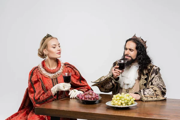 Reina rubia mirando al rey hispano en ropa medieval oliendo vino tinto en vidrio aislado en blanco - foto de stock