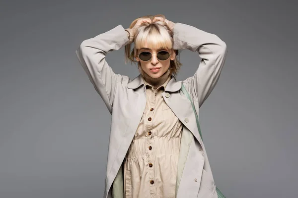 Young woman in sunglasses and trench coat posing while looking at camera isolated on grey — Stock Photo
