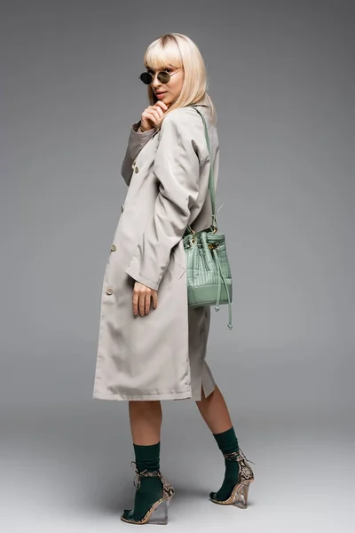 Full length of young woman in sunglasses and trench coat with green bag standing on grey — Stock Photo