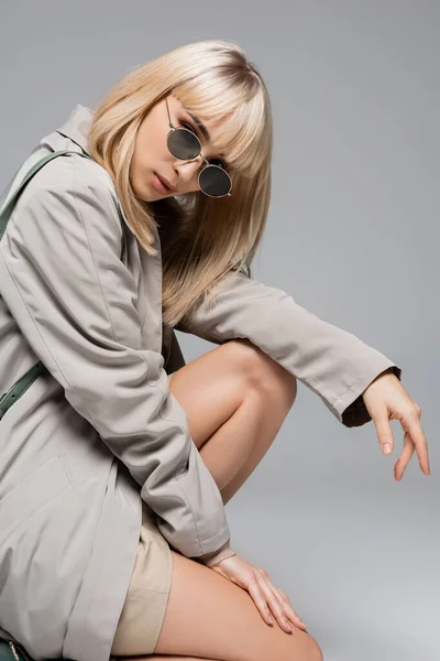 Jeune femme à la mode en lunettes de soleil et trench coat regardant la caméra isolée sur gris — Photo de stock