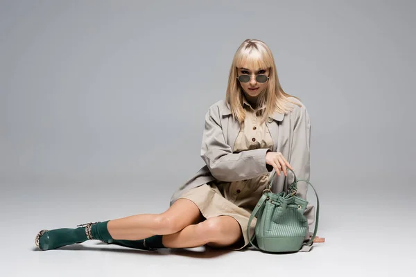 Full length of trendy young woman in sunglasses and trench coat posing near bag on grey — Stock Photo