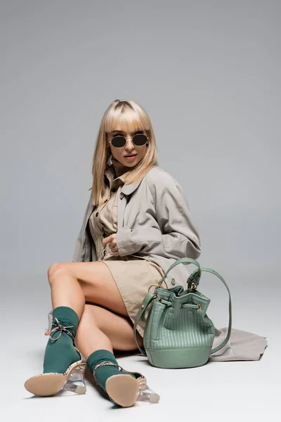 Full length of trendy woman in sunglasses and trench coat posing with bag on grey — Stock Photo