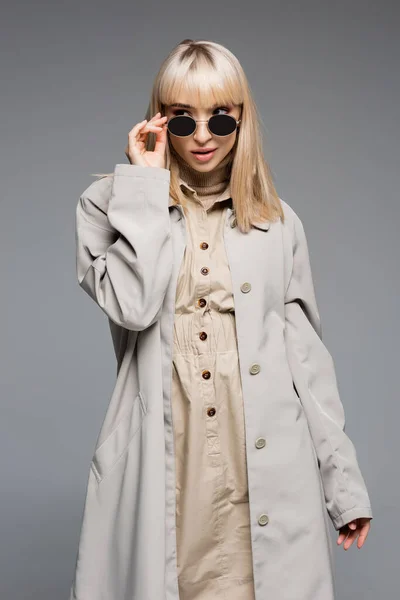 Модна молода жінка в тренч пальто, що регулює сонцезахисні окуляри ізольовані на сірому — стокове фото