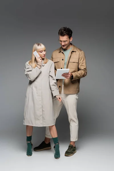 Longitud completa del modelo de moda hablando en el teléfono inteligente cerca de hombre con estilo con la tableta digital de pie en gris — Stock Photo