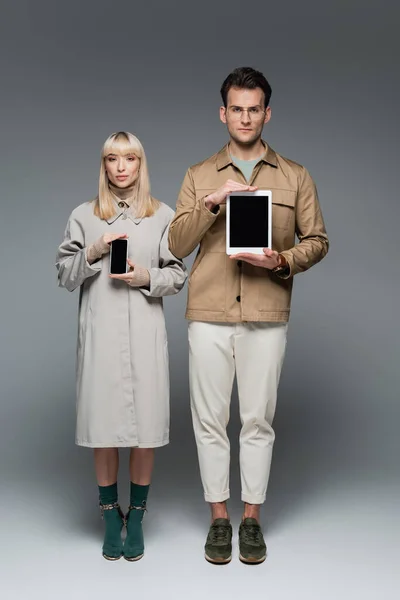 Full length of trendy models posing while holding gadgets with blank screen on grey — Stock Photo