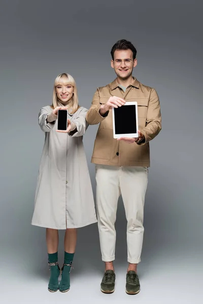Full length of happy trendy models holding gadgets with blank screen on grey — Stock Photo