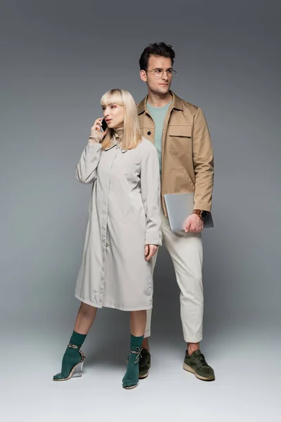 Longitud completa del modelo de moda hablando en el teléfono inteligente cerca de hombre elegante celebración de la computadora portátil en gris — Stock Photo