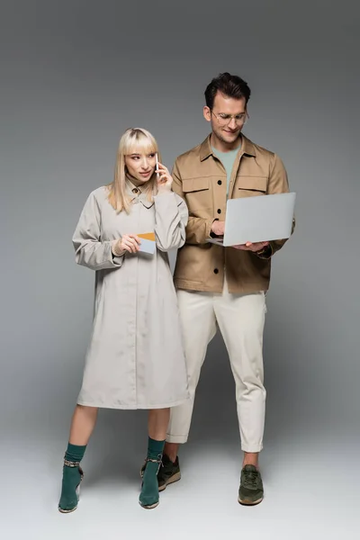 Young woman in shirt dress talking on smartphone and holding credit cards near stylish man using laptop on grey — Stock Photo