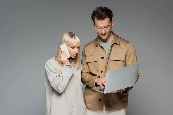 Trendy model talking on smartphone near stylish man using laptop isolated on grey — Stock Photo