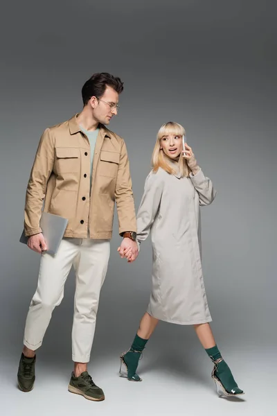 Longitud completa de la mujer joven en vestido de camisa hablando en el teléfono inteligente y tomados de la mano con el hombre elegante caminando con el ordenador portátil en gris - foto de stock