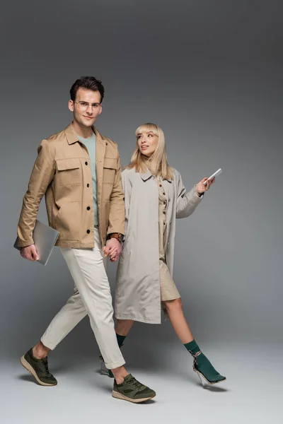 Full length of trendy couple holding hands while walking with gadgets on grey — Stock Photo