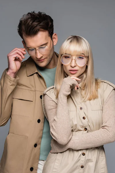 Mujer joven con estilo mirando a la cámara cerca del hombre ajustando gafas aisladas en gris - foto de stock