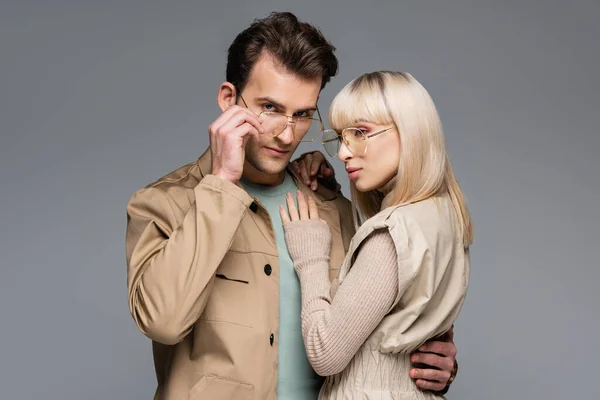 Стильная пара в очках, обнимающая, позируя изолированной на сером — стоковое фото