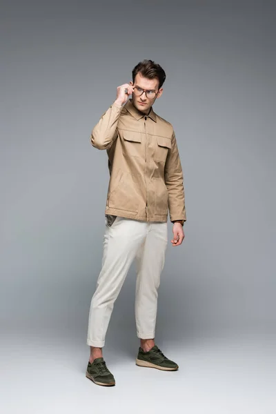 Full length of trendy man in stylish outfit posing while adjusting glasses on grey — Stock Photo