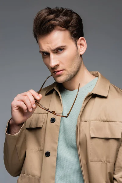 Uomo alla moda in giacca tenendo gli occhiali isolati su grigio — Foto stock