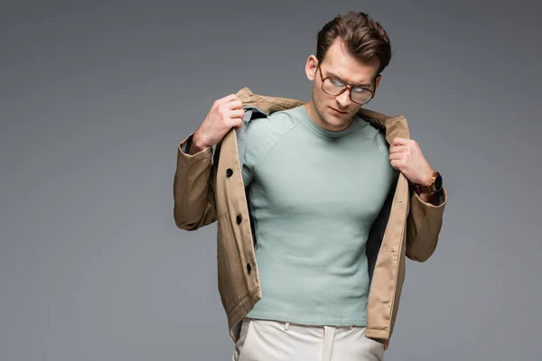 Модний чоловік в окулярах регулюючи куртку, позує ізольовано на сірому — стокове фото