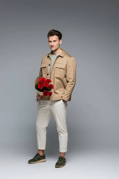 In voller Länge trendiger Mann in stylischem Outfit mit Rosen und einem Lächeln auf grau — Stockfoto