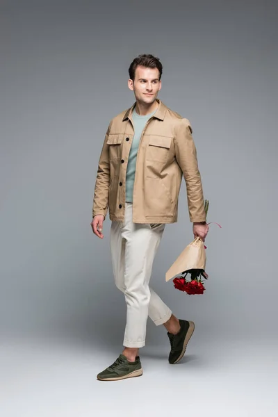 Full length of trendy man holding bouquet of red roses wrapped in paper on grey — Stock Photo