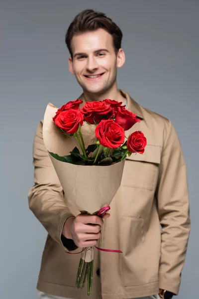 Hombre sonriente borroso sosteniendo ramo de rosas rojas aisladas en gris - foto de stock