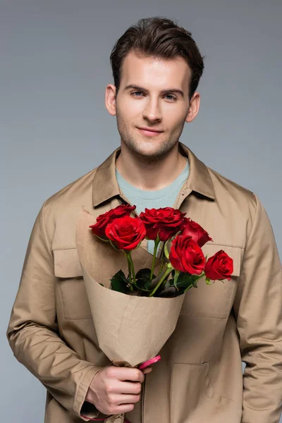 Sonriente hombre sosteniendo ramo de rosas rojas aisladas en gris - foto de stock