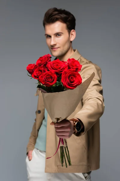 Homme flou à la mode tenant bouquet de roses rouges enveloppé dans du papier isolé sur gris — Photo de stock