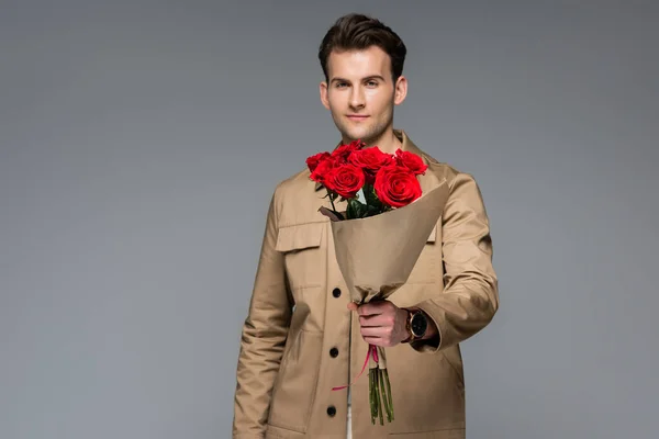 Hombre de moda sosteniendo ramo de rosas rojas envueltas en papel aislado en gris - foto de stock