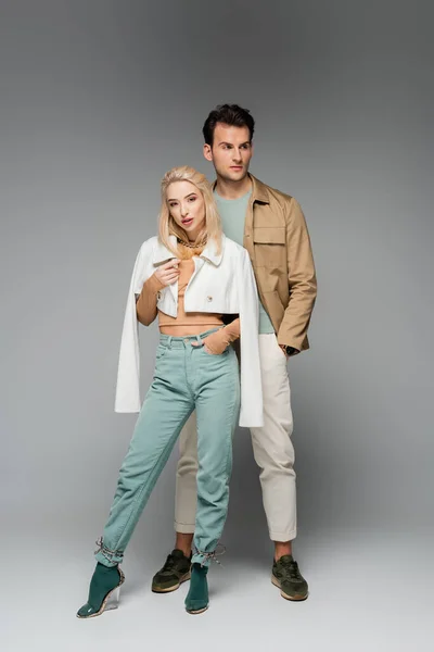 Full length of trendy models in pants and jackets posing with hands in pockets on grey — Stock Photo