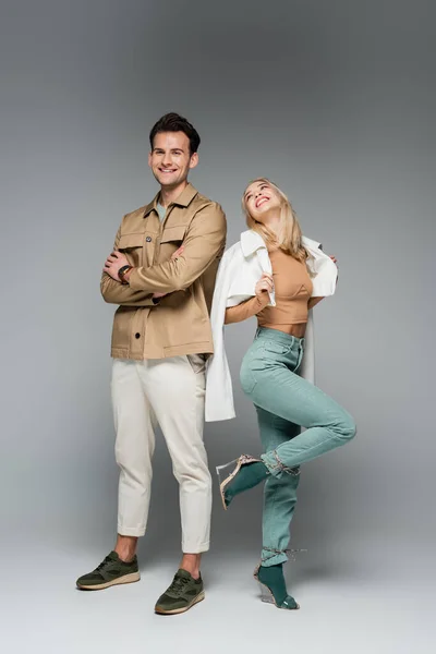 Longitud completa de modelos de moda en pantalones y chaquetas posando y sonriendo en gris - foto de stock